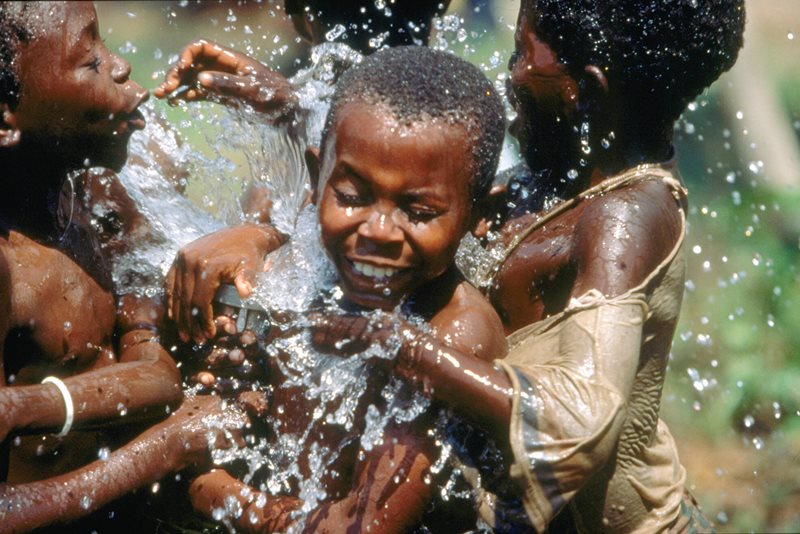 Tre stycken unga pojkar badar i vatten, som finns vid ett av ICRCs vattendistributionsprojekt. Man kan inte se vattnets ursprungskälla, utan vattnet sprutar upp mellan barnen. Bilden utstrålar glädje.