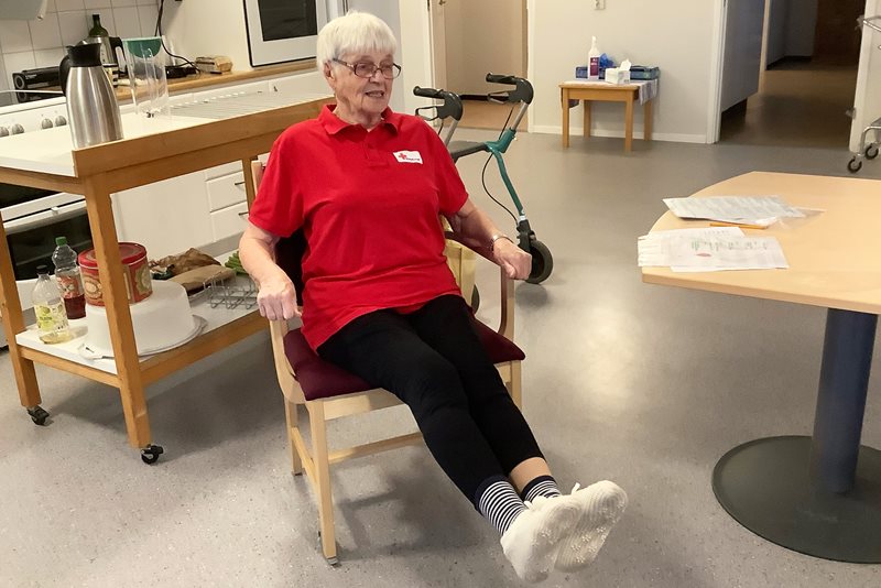 Vivi 88 år sitter på en stol och visar rörelser för sittgympan. Hennes ben är rakt utsträckta. 